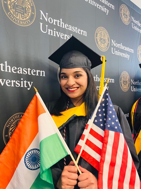Photo of Preeti Jain in her graduation regalia, holding one U.S. flag and one India flag and smiling in front of a backdrop with the Northeastern seal