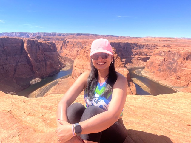 A photo of Preeti Jain sitting at the rim of the Grand Canyon in sunglasses and a hat, smiling