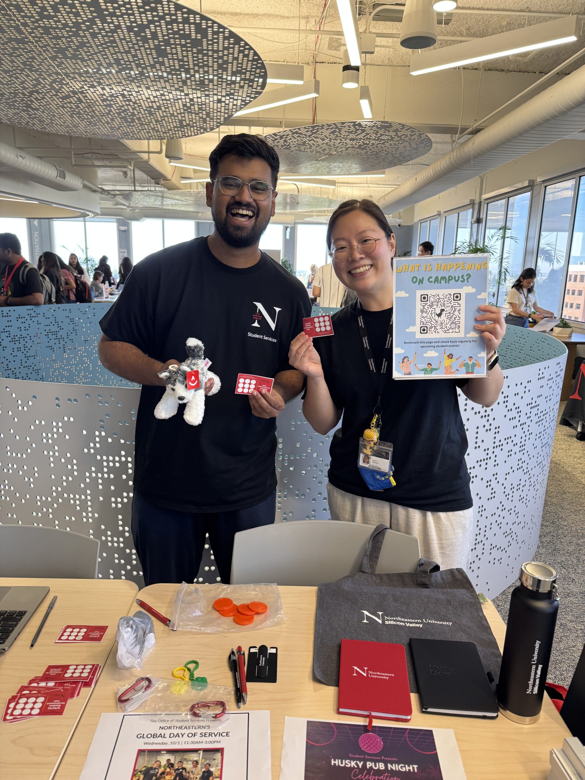 A photo of a staff member and a student ambassador smiling and holding up some Northeastern gear at an event on campus