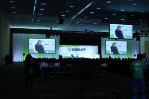 A shot from the back of the audience showing a woman being interviewed on stage with several large screens showing a closeup of her as she answers.
