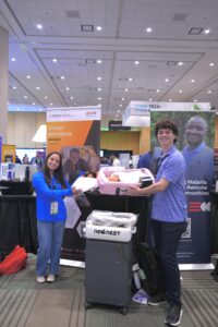 Members of the Che Innovations team posing with their product, the NeoNest. A young man is holding a pink rectangular bassinet with a baby doll inside, and behind him a woman holds another similar item.