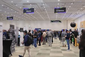 A view of a crowded room in the Moscone Center with signs on the ceiling for different sectors like "Health", "Enterprise", "Consumer"
