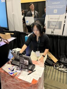 A photo of Yi seated at a demo booth, looking intently at a screen in front of her with headphones on.