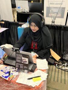 A photo of Aisha trying out a demo at a station; she is wearing over ear headphones and looking at a screen in front of her. A sign reads "Multi-Spectral Reality Detector"