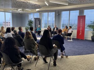 A view from the audience as the panel discussion is held; the panelists are seated in front of tall windows looking out over San Jose