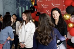 A group of alumni laugh together at a mixer; in the background are balloons and a large Northeastern step and repeat banner