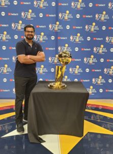 Sahil Gothoskar stands on a basketball court in front of a step & repeat backdrop with the logos of the NBA Finals, abc, and YouTube. He is crossing his arms and smiling next to a table where the trophy is placed on top of a black tablecloth.
