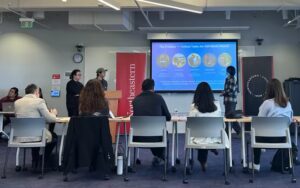 Three young men stand at the front of the room, two at a lectern at the side while the third stands in front of the projection screen. He gestures at a blue slide with illustrations. The judges are seen in the foreground.