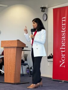 Holding a microphone and dressed in a white blazer, the lead judge speaks with a smile and holds up a finger while making a point.