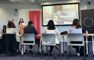 Standing at the lectern next to a screen showing an image of a living room with the name of the proposed furnishing app, Ottera, a student pauses and makes an expression of contemplation after hearing a question from the judges.