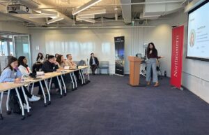 A row of judges is seen listening to the presentation, some taking notes, as the student gestures to her deck and speaks