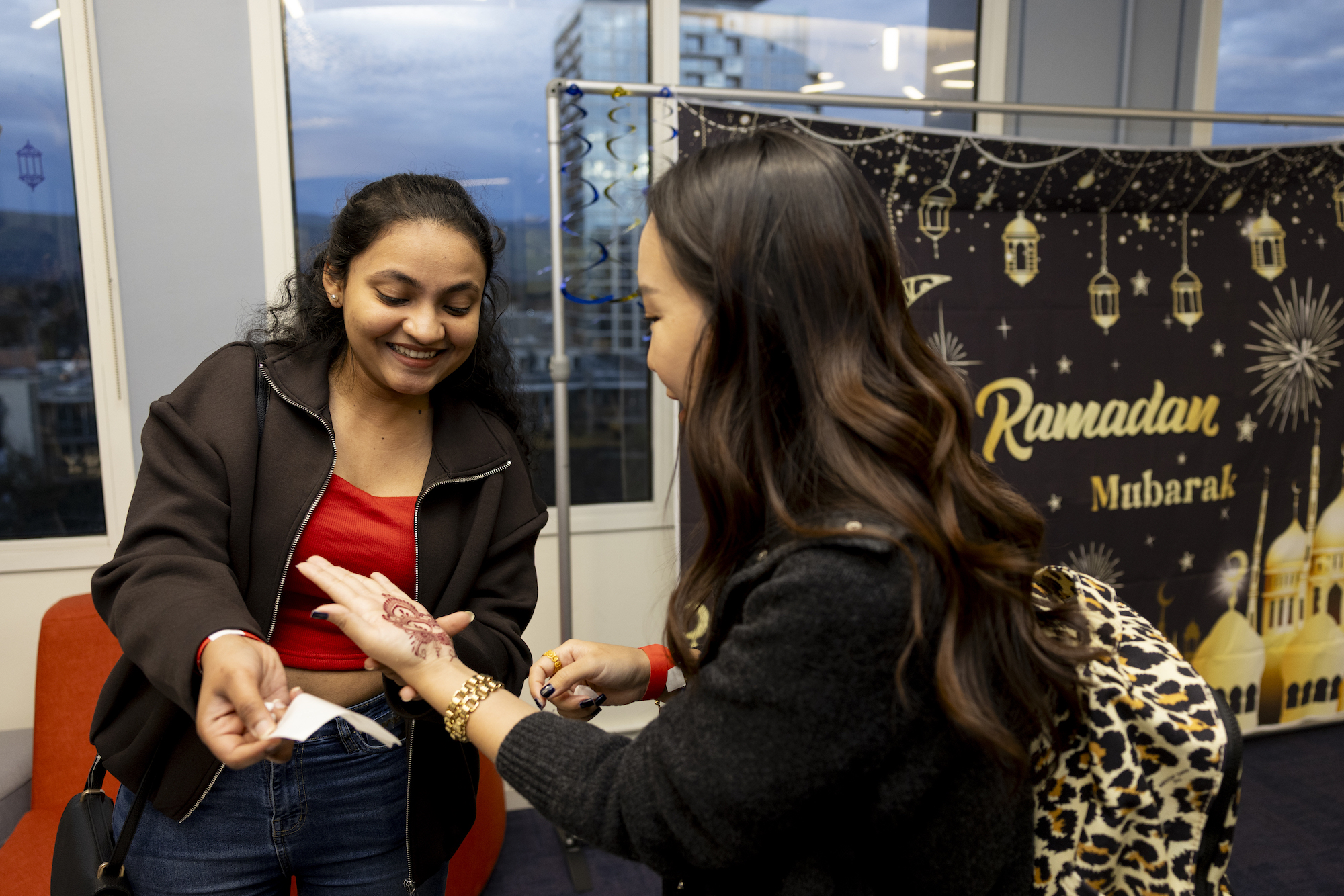 One woman holds the hand of another as she pulls off the backing of a temporary henna tattoo, both smiling; in the background a Ramadan Mubarak photo backdrop can be seen