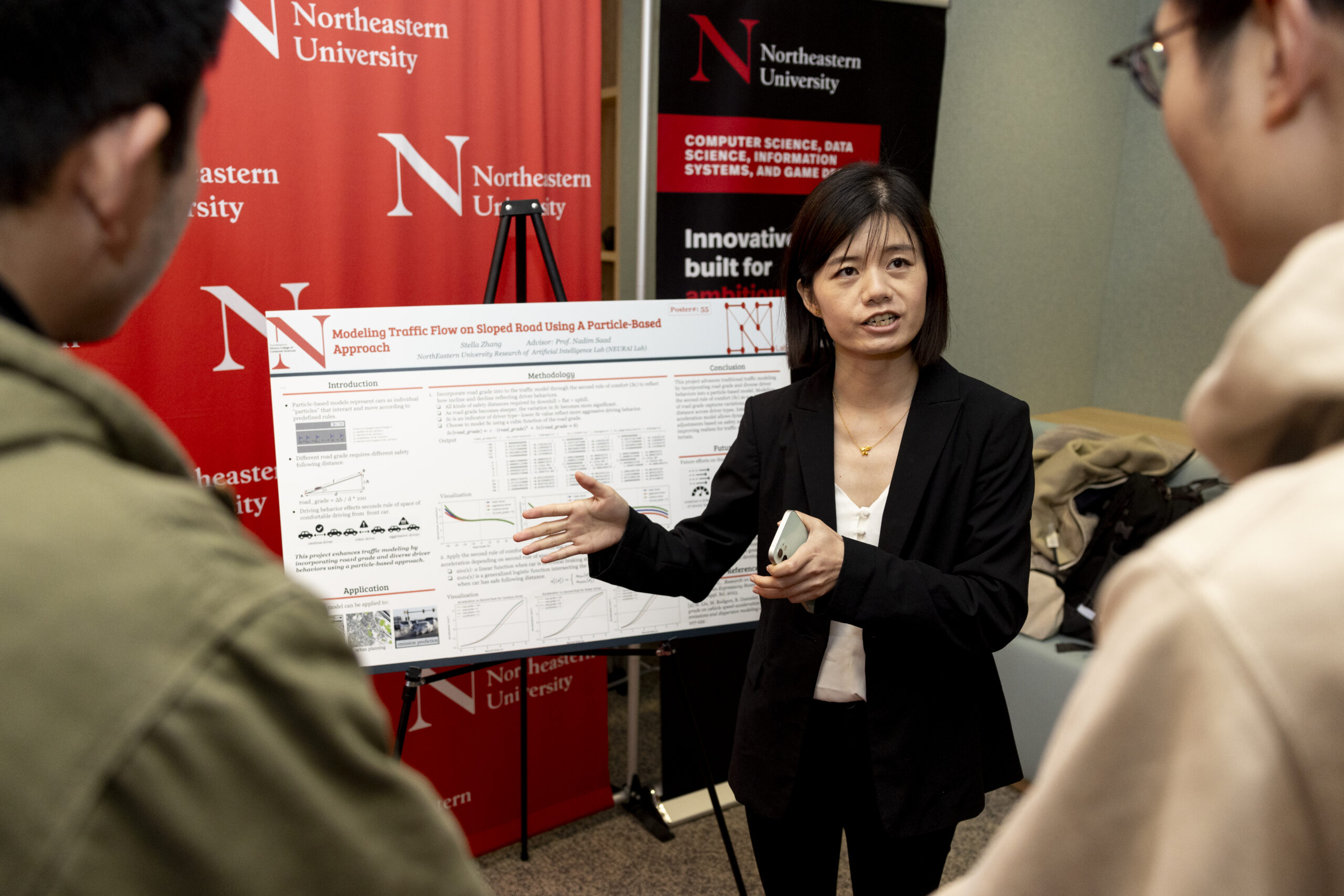 A student stands in front of her research poster, speaking to two people who are standing in the foreground listening
