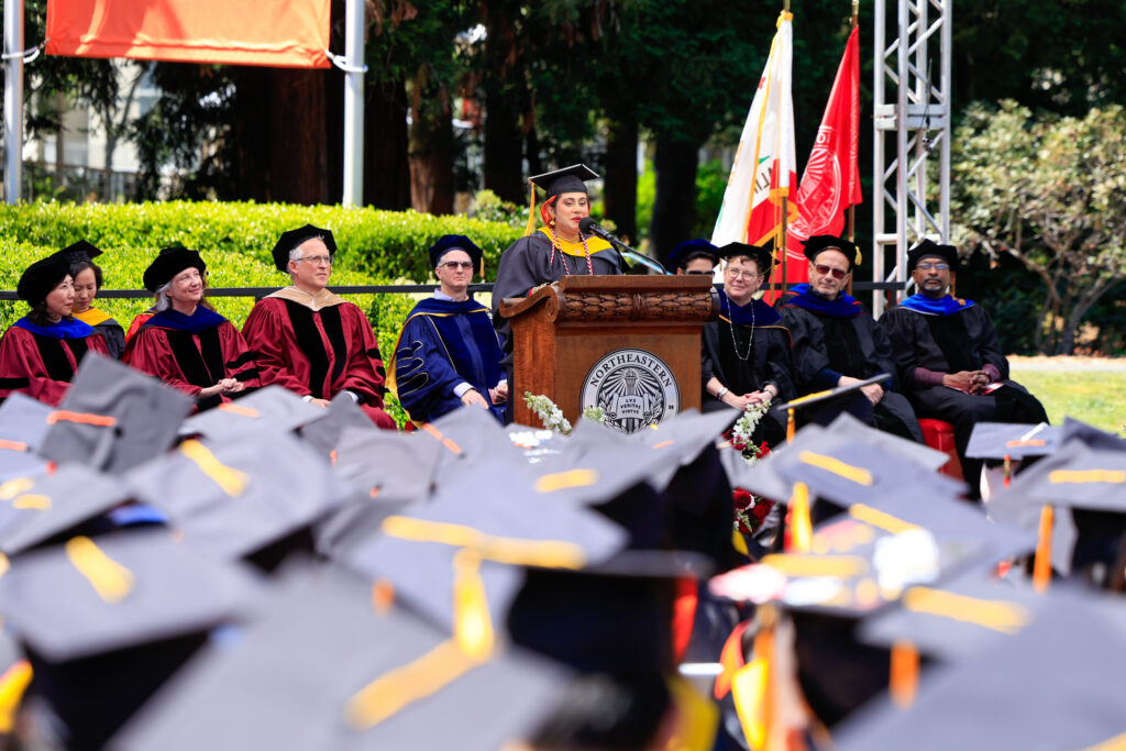 Aisha Abdur Rahim speaks at the podium, with a field of graduation caps visible in the foreground