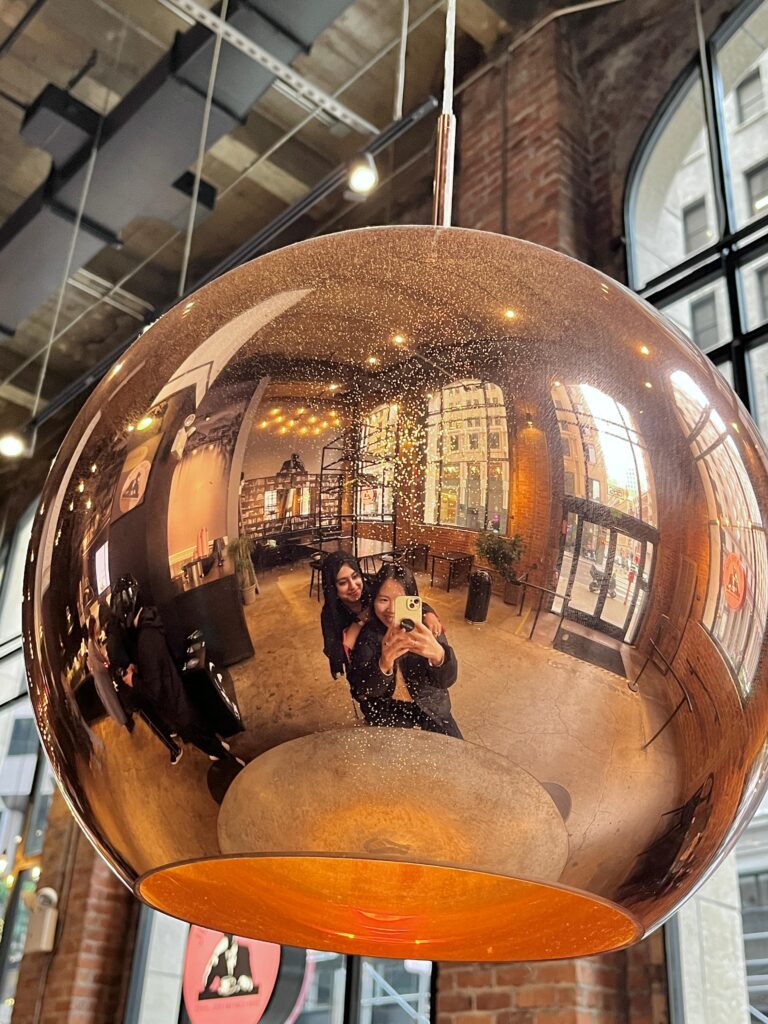 A photo of a reflective copper light fixture hanging from the ceiling in a cafe. Reflected are Yi and Aisha, who are posing for a photo