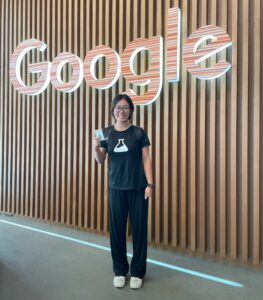 Wearing a Google Labs t-shirt and holding up her employee badge, Sonting poses in front of the Google sign in the lobby of her building at work.