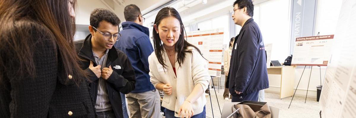 A student researcher points towards her project while explaining her work to a group of peers who lean in to listen.